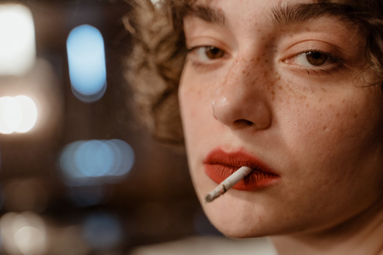 our-story Close-up portrait of a young woman with red lips and freckles holding a cigarette.